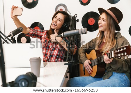 Happy woman singer performing at radio program while making podcast recording for online show, playing guitar, taking selfie with a host