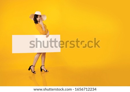 Young brunette woman with pin-up hairstyle in a yellow bathing suit and white hat holds a white poster in her hands and poses on a yellow background