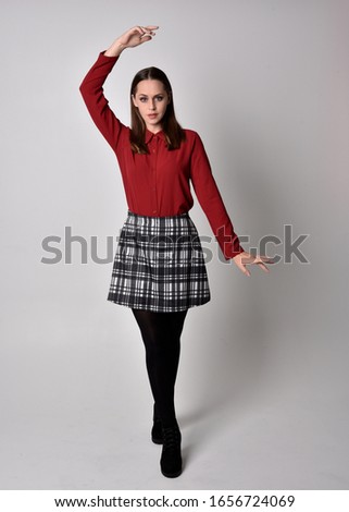 full length portrait of a pretty brunette girl wearing a red shirt and plaid skirt with leggings and boots. Standing pose with hand gesture against a  studio background.
