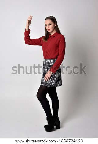 full length portrait of a pretty brunette girl wearing a red shirt and plaid skirt with leggings and boots. Standing pose with hand gesture against a  studio background.

