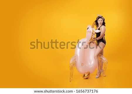 Young brunette woman with pin-up hairstyle in a black swimsuit posing with a beach inflatable unicorn toy on a yellow background