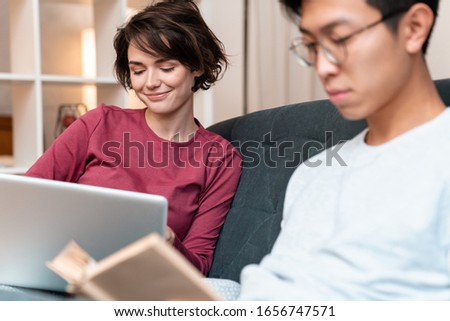 Photo of pleased multinational couple reading book and using laptop while sitting on sofa at home