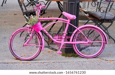 A pink coloured old, retro, vintage bicycle with a basket of flowers leaning against a light post.