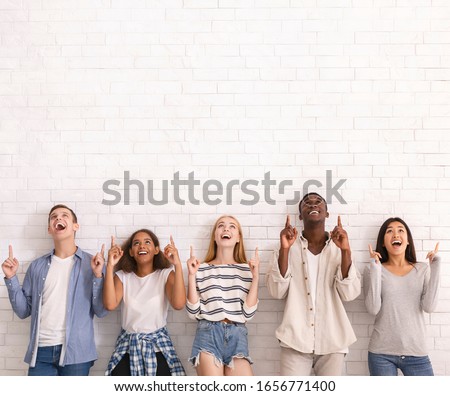 Wow, look at that. Group of multiracial excited friends pointing up at copy space over white brick wall