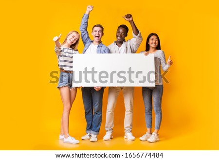 Excited international group of teenagers holding empty white plackard, yellow background