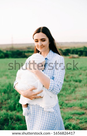 portrait of a young mother in a blue checked dress, holding a small child to her chest, in the rays of the setting summer sun