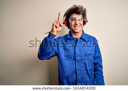 Young constructor man wearing uniform and earmuffs over isolated white background pointing finger up with successful idea. Exited and happy. Number one.