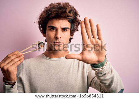 Young handsome man eating sushi using chopsticks over isolated pink background with open hand doing stop sign with serious and confident expression, defense gesture
