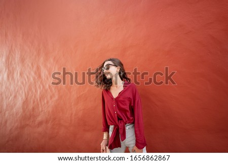 Portrait of an active young woman dressed in bright shirt on the red wall background outdoors. Carefree lifestyle and women's fashion concept