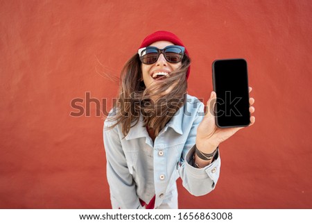 Young stylish woman showing mobile phone while standing relaxed on the red wall background. Phone with black screen to copy paste