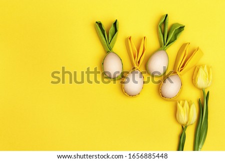 Funny Easter concept. Yellow flowers tulip and wooden easter egg with fabric ears as fun bunny on yellow background. Flat lay, top view.