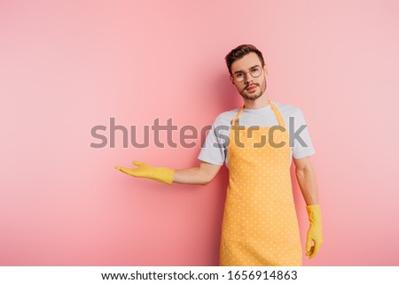 serious young man in apron and rubber gloves standing with open arm on pink background