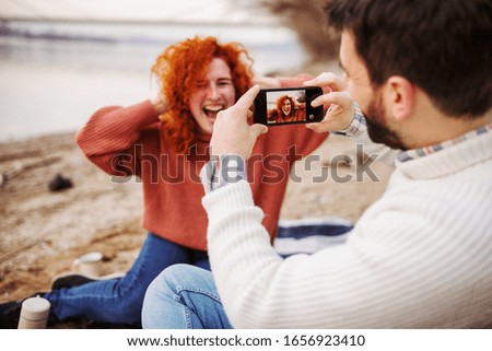 Young happy couple sitting by the river and having fun. a young guy takes a picture of his girlfriend on the phone while sitting on a picnic by the river
