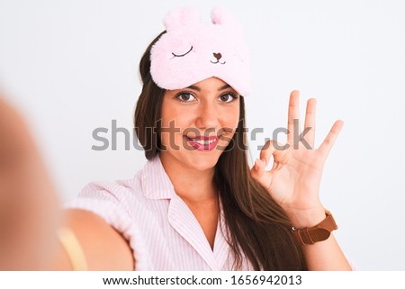 Girl wearing pajama and sleep mask make selfie by camera over isolated white background doing ok sign with fingers, excellent symbol