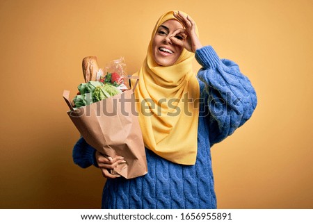 Young beautiful brunette arab woman wearing islamic hijab holding paper bag with food with happy face smiling doing ok sign with hand on eye looking through fingers