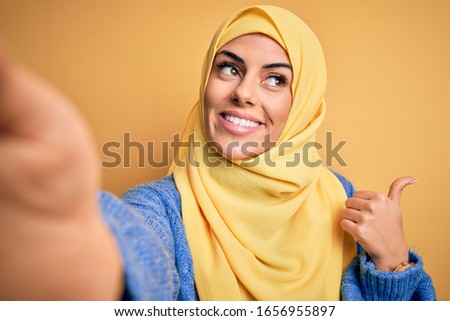 Young beautiful brunette arab woman wearing islamic hijab make selfie by camera pointing and showing with thumb up to the side with happy face smiling