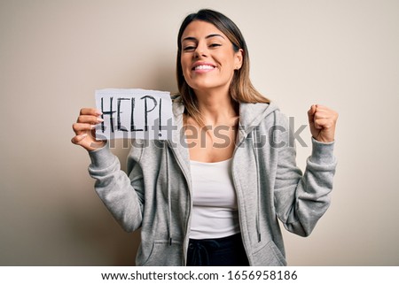 Young beautiful brunette woman holding paper with help message over white background screaming proud and celebrating victory and success very excited, cheering emotion