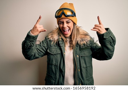 Young brunette skier woman wearing snow clothes and ski goggles over white background shouting with crazy expression doing rock symbol with hands up. Music star. Heavy music concept.
