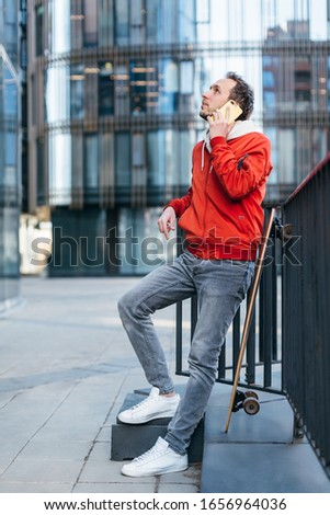 Fashionable man in red jacket, jeans and white sneakers talking on the mobile phone. Longboard is propped up next to it. Selective focus, blurred background. Urban and street photo. Concept of leisure