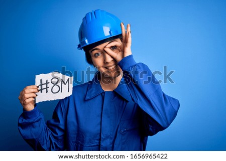 Beautiful worker woman wearing hardhat and uniform celebrating 8th march womens day with happy face smiling doing ok sign with hand on eye looking through fingers