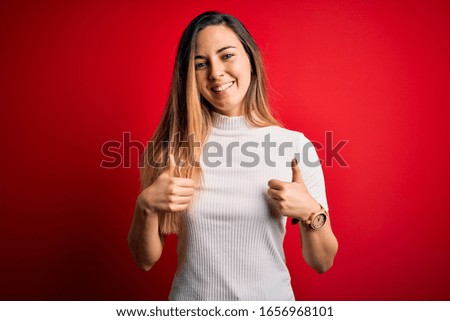 Beautiful blonde woman with blue eyes wearing casual white t-shirt over red background success sign doing positive gesture with hand, thumbs up smiling and happy. Cheerful expression 
