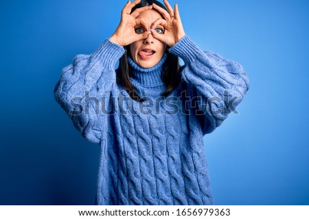 Young brunette woman with blue eyes wearing casual turtleneck sweater doing ok gesture like binoculars sticking tongue out, eyes looking through fingers. Crazy expression.