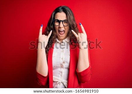 Young beautiful woman with curly hair wearing jacket and glasses over red background shouting with crazy expression doing rock symbol with hands up. Music star. Heavy concept.