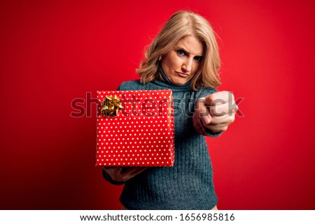 Middle age beautiful blonde woman holding birthday gift over isolated red background annoyed and frustrated shouting with anger, crazy and yelling with raised hand, anger concept