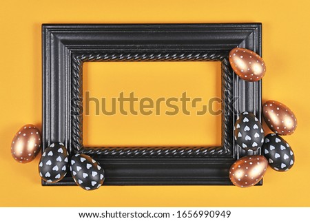 Easter design with blank black elegant picture frame surrounded by modern golden, black and white easter eggs on yellow background
