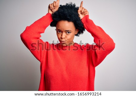 Young beautiful African American afro woman with curly hair wearing red casual sweater doing funny gesture with finger over head as bull horns