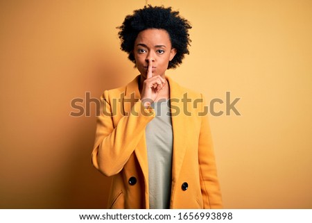 Young beautiful African American afro businesswoman with curly hair wearing yellow jacket asking to be quiet with finger on lips. Silence and secret concept.