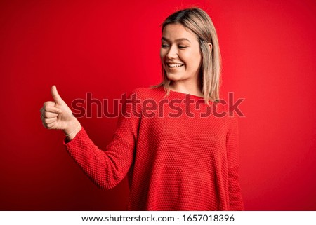 Young beautiful blonde woman wearing casual sweater over red isolated background Looking proud, smiling doing thumbs up gesture to the side