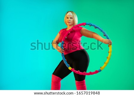 Young caucasian plus size female model's training on gradient green background in neon light. Doing workout exercises with the hoop. Concept of sport, healthy lifestyle, body positive, equality.