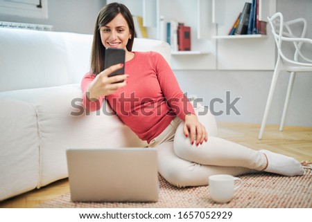 Young attractive smiling woman sitting on the floor at home, taking selfie and relaxing.