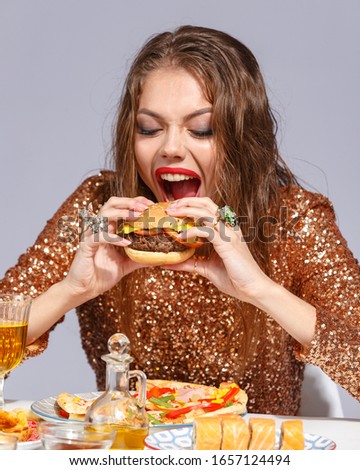 Beautiful young girl with bright makeup in an evening dress with an appetite eats fast food strength at the table. Creative fast food concept