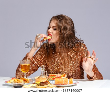 Beautiful young girl with bright makeup in an evening dress with an appetite eats fast food strength at the table. Creative fast food concept
