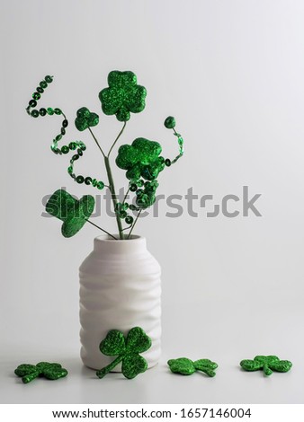St. Patrick’s Day green swirls, four leaf clovers and a hat in a white vase with more glittery green four leaf clovers on a white background.