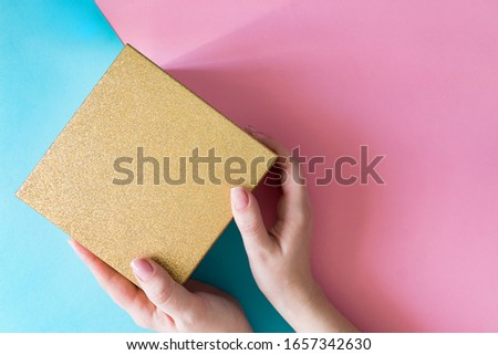 Woman holding gold sparkling gift box on color blue and pink background. Festive greeting card. Holiday concept