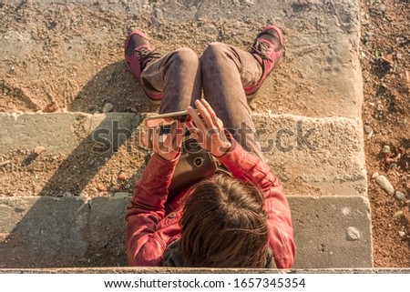 Top view of a brunette woman sitting on the stairs of a beach, shooting a photogragh or video with her mobile phone on a sunny autumn day.