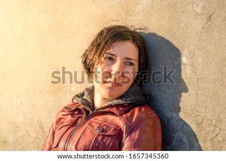 Brunette woman with red leather jacket leaning to a gray concrete wall sitting alone under the sunlight. Sadness and loneliness concept.