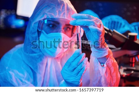 Nurse in mask and white uniform, holding tube with liquid and sitting in neon lighted laboratory with computer and medical equipment searching for Coronavirus vaccine.