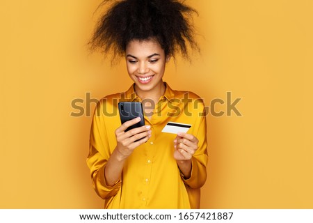 Young girl makes a payment, using a credit card and smartphone. Photo of african american girl on yellow background