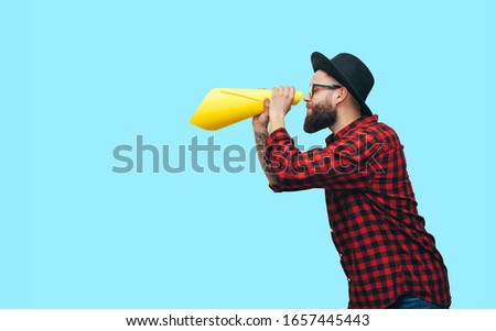 Handsome hipster guy wearing a red plaid shirt and hat looking through yellow rolled up paper as telescope. Your text here! Discount, sale, season sales.