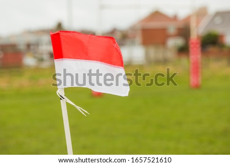 Red and white football or rugby flag on a white pole infant of a grass pitch with stands behind. blowing in the wind. English stormy winds but games still happening with the flag going quickly 