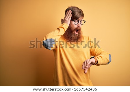 Handsome Irish redhead man with beard wearing glasses over yellow isolated background Looking at the watch time worried, afraid of getting late