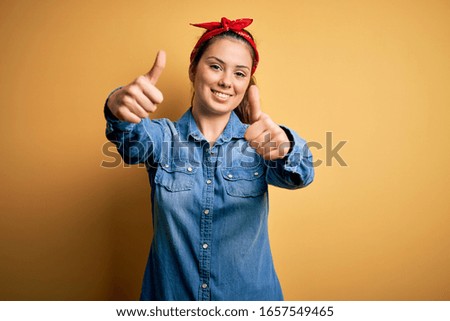 Young beautiful brunette woman wearing casual denim shirt and hair handkerchief approving doing positive gesture with hand, thumbs up smiling and happy for success. Winner gesture.