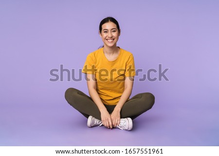 Smiling pretty young woman sitting on a floor with legs crossed isolated over violet background, looking at camera