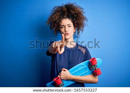 Young sporty woman with curly hair and piercing holding skate over blue background pointing with finger to the camera and to you, hand sign, positive and confident gesture from the front