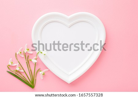 Frame of white heart shape and fresh snowdrops on light pink table background. Pastel color. Love and happiness concept. Empty place for cute, emotional, sentimental text, quote or sayings. Closeup.