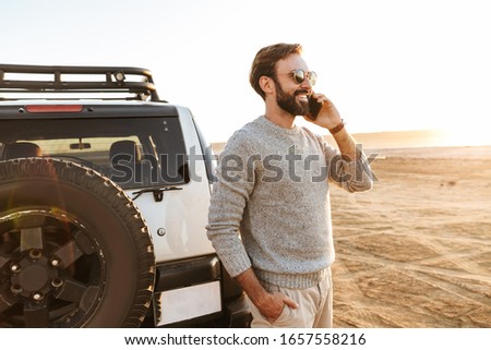 Smiling handsome young man using mobile phone while standing at the car at the sunny beach, calling